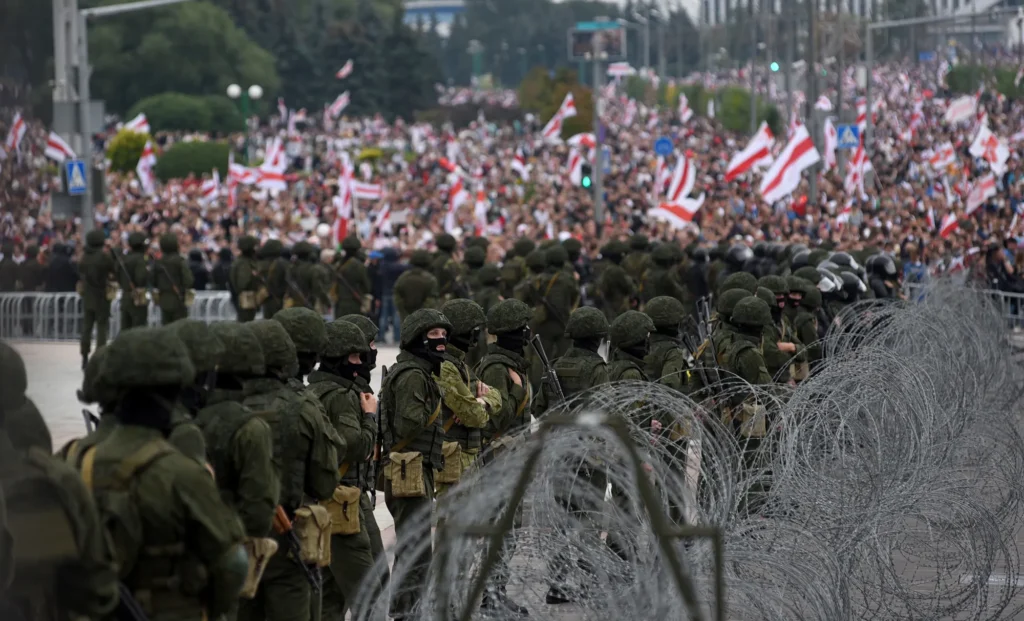 Heavily armed Belarusian soldiers stand in formation behind barbed wire, blocking a massive crowd waving historic red-and-white flags, symbolising state repression and control during the Belarus cybersecurity crackdown in 2025.