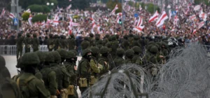 Heavily armed Belarusian soldiers stand in formation behind barbed wire, blocking a massive crowd waving historic red-and-white flags, symbolising state repression and control during the Belarus cybersecurity crackdown in 2025.