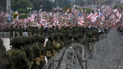 Heavily armed Belarusian soldiers stand in formation behind barbed wire, blocking a massive crowd waving historic red-and-white flags, symbolising state repression and control during the Belarus cybersecurity crackdown in 2025.