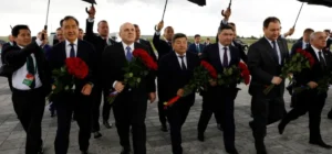 Belarus economic crisis illustrated by officials in suits holding red roses under umbrellas during a formal ceremony, symbolising political control amid sanctions and mismanagement.