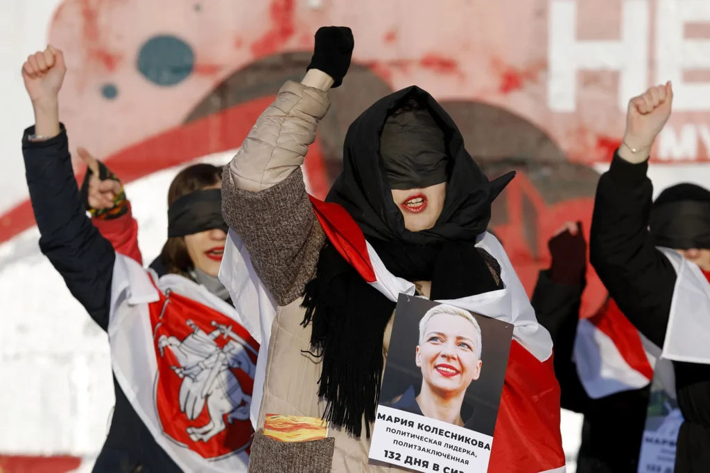 Women protesting in Belarus with eyes covered, holding a portrait of political prisoner Maria Kolesnikova, wrapped in the historical white-red-white flag — a powerful example of Belarus resistance art against state repression.