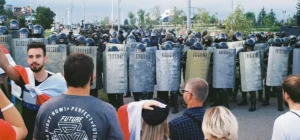 Belarus state media protests in 2025, showing demonstrators wrapped in white-red-white flags confronting heavily armed riot police with shields in Minsk.