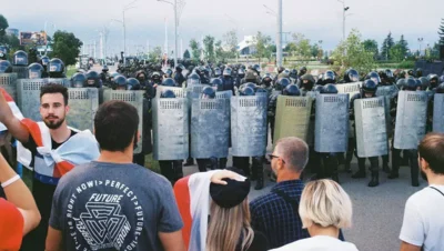 Belarus state media protests in 2025, showing demonstrators wrapped in white-red-white flags confronting heavily armed riot police with shields in Minsk.