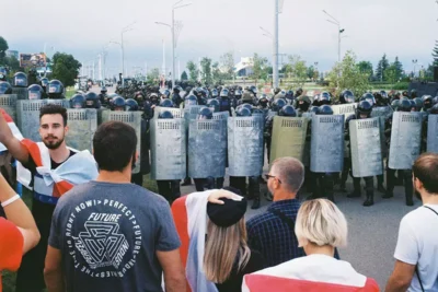Belarus state media protests in 2025, showing demonstrators wrapped in white-red-white flags confronting heavily armed riot police with shields in Minsk.