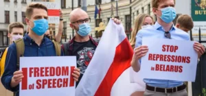 Belarus youth movements activists holding protest signs reading 'Freedom of Speech' and 'Stop Repressions in Belarus' while wearing face masks and carrying the historic white-red-white flag during a demonstration.