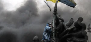Protester in protective gear climbing a monument and waving the Ukrainian flag amid thick smoke during the conflict, symbolising Belarus’s role in Russia’s Ukraine war in 2025.