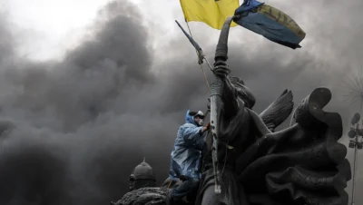 Protester in protective gear climbing a monument and waving the Ukrainian flag amid thick smoke during the conflict, symbolising Belarus’s role in Russia’s Ukraine war in 2025.