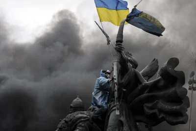 Protester in protective gear climbing a monument and waving the Ukrainian flag amid thick smoke during the conflict, symbolising Belarus’s role in Russia’s Ukraine war in 2025.