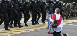 Belarus human rights violations 2025 – Woman draped in Belarus opposition flag kneels before riot police during a peaceful protest, symbolising defiance against state repression.