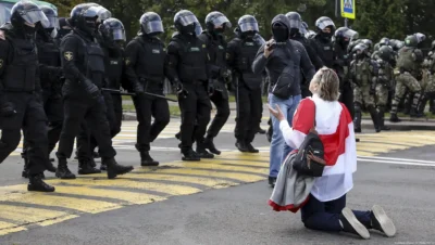 Belarus human rights violations 2025 – Woman draped in Belarus opposition flag kneels before riot police during a peaceful protest, symbolising defiance against state repression.