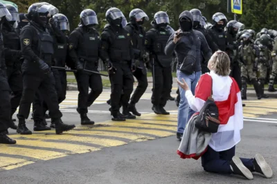 Belarus human rights violations 2025 – Woman draped in Belarus opposition flag kneels before riot police during a peaceful protest, symbolising defiance against state repression.