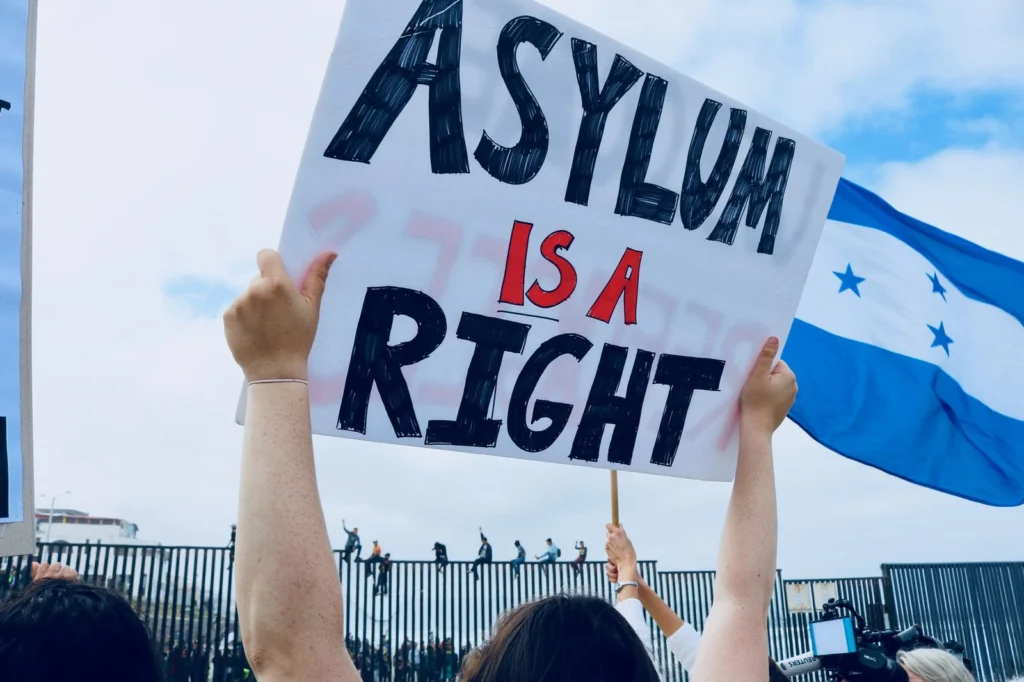 Protester holding a sign that reads “Asylum is a Right” in front of a border fence, with a Honduran flag visible—symbolising global demands for Political Asylum and human rights.
